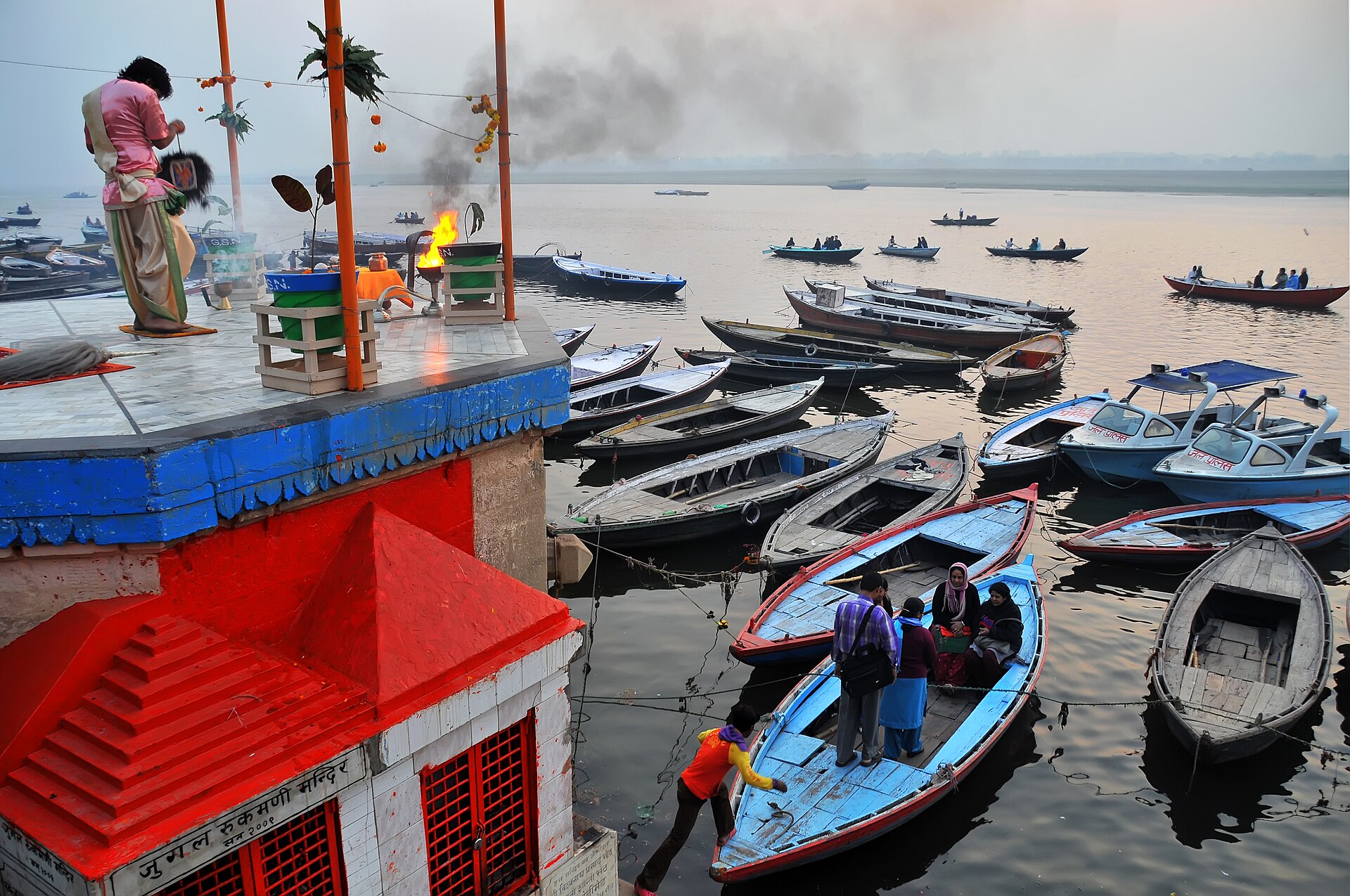 Boat rides — wooden boats moored below the ghat at dawn, pilgrims stepping in, mist on the Ganga