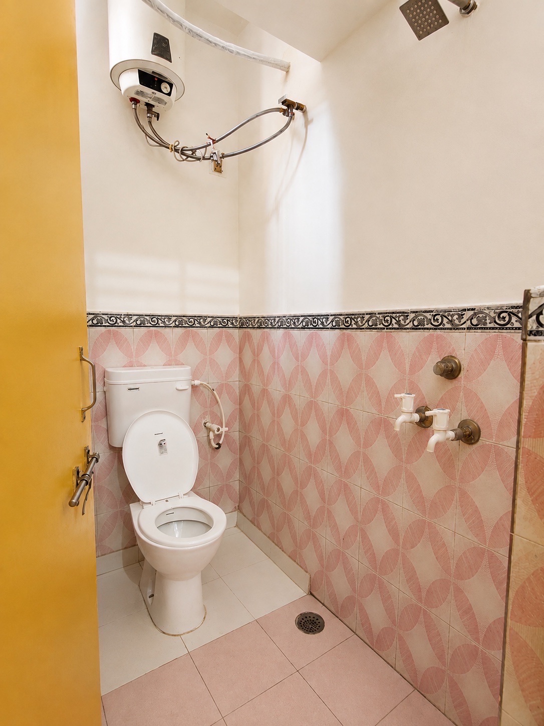 Gardenia, Varanasi: second bathroom, overhead water heater, shower and mixer taps on pink patterned tiling, yellow door with a towel rail