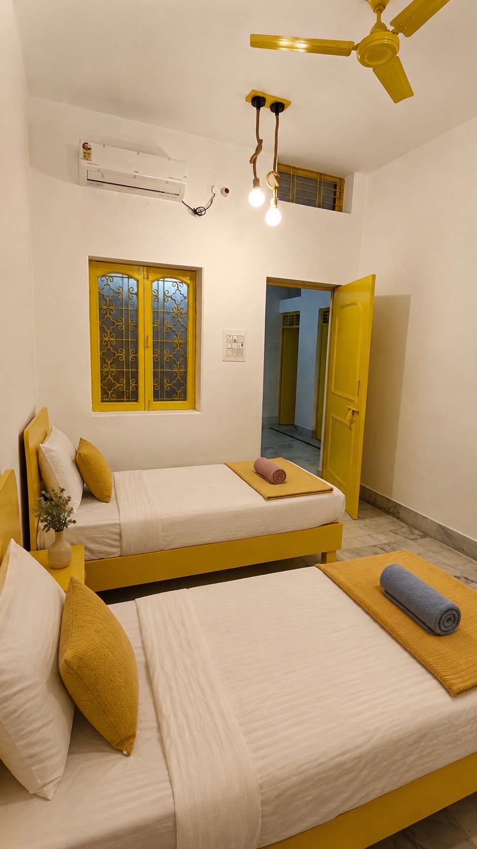 Gardenia, Varanasi: twin beds under a yellow ceiling fan and air conditioning, a grilled yellow window between them and the door open to the hall