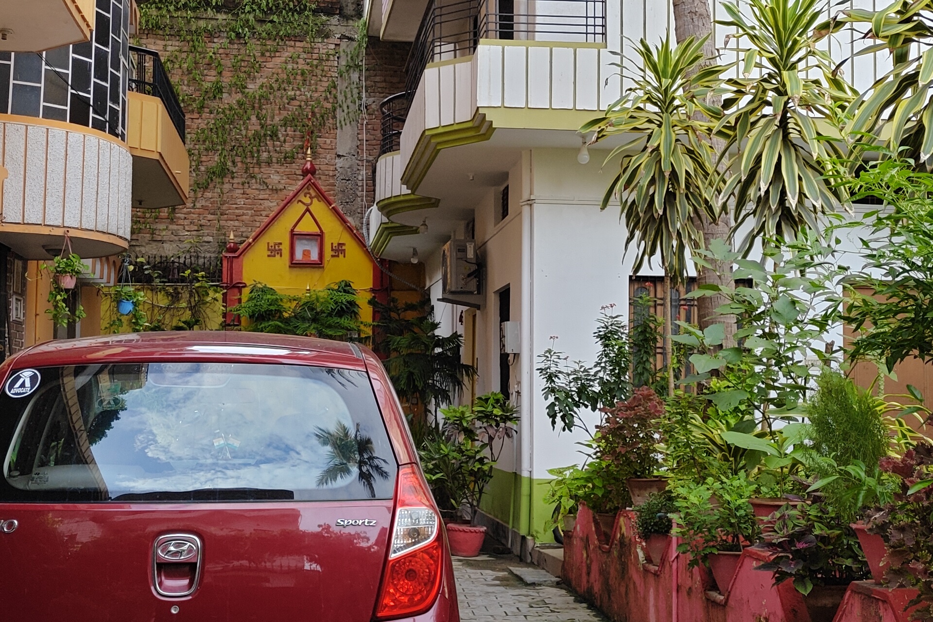 Gardenia, Varanasi: a car parked beside the house, a small yellow and red wayside shrine on the wall, balconies and palms above