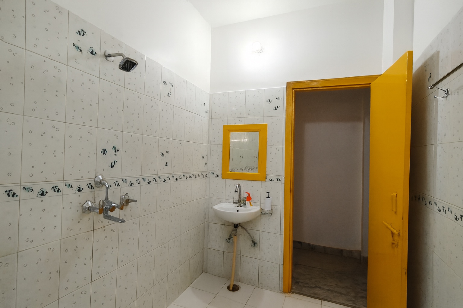 Minimilist, Varanasi: second bathroom, wall basin under a yellow-framed mirror, shower and mixer taps on the tiled wall, saffron door open to the room beyond