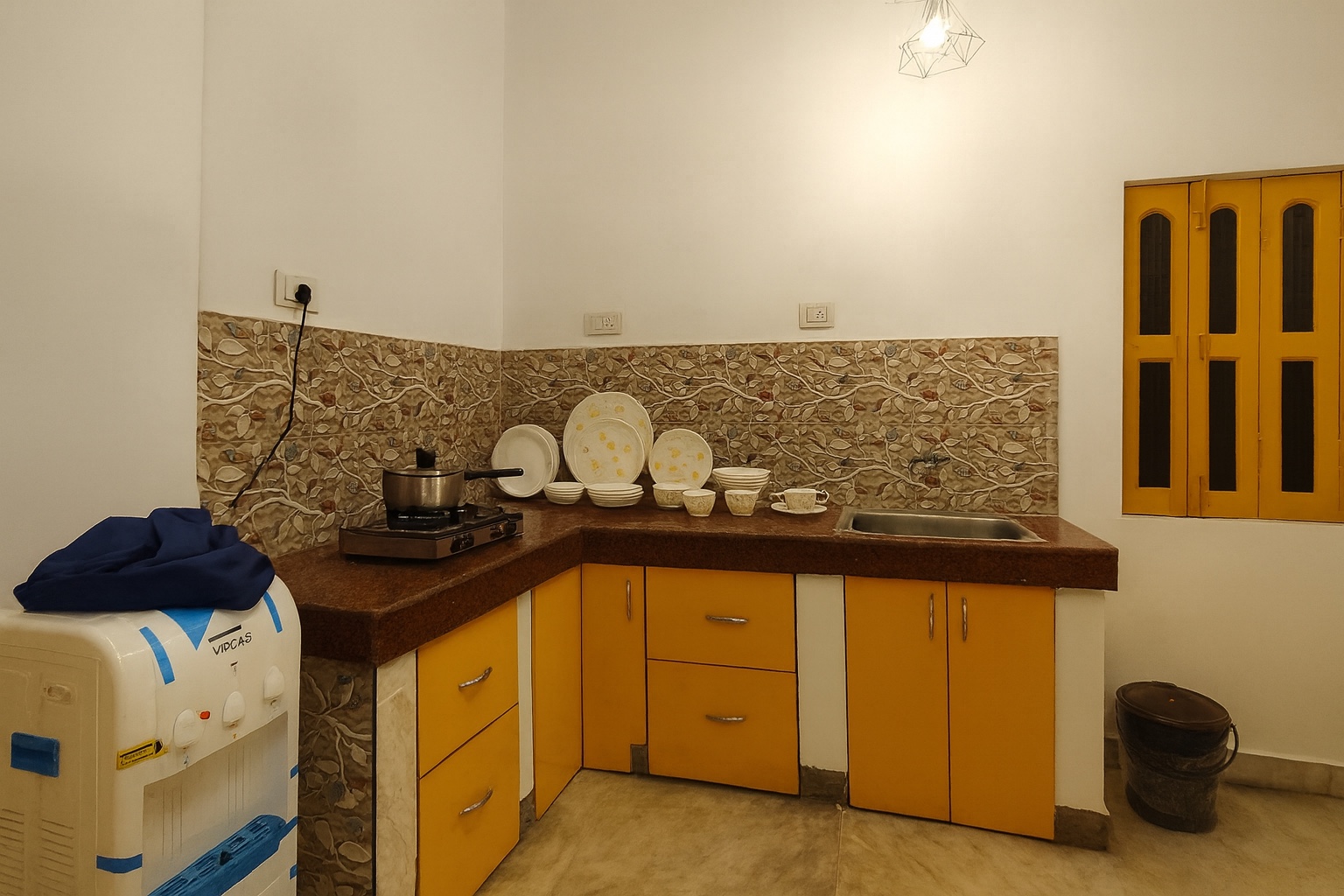 Minimilist, Varanasi: L-shaped granite counter with mustard cabinets, a single gas burner with a steel pan, stacked white crockery and a water cooler under a yellow louvred window