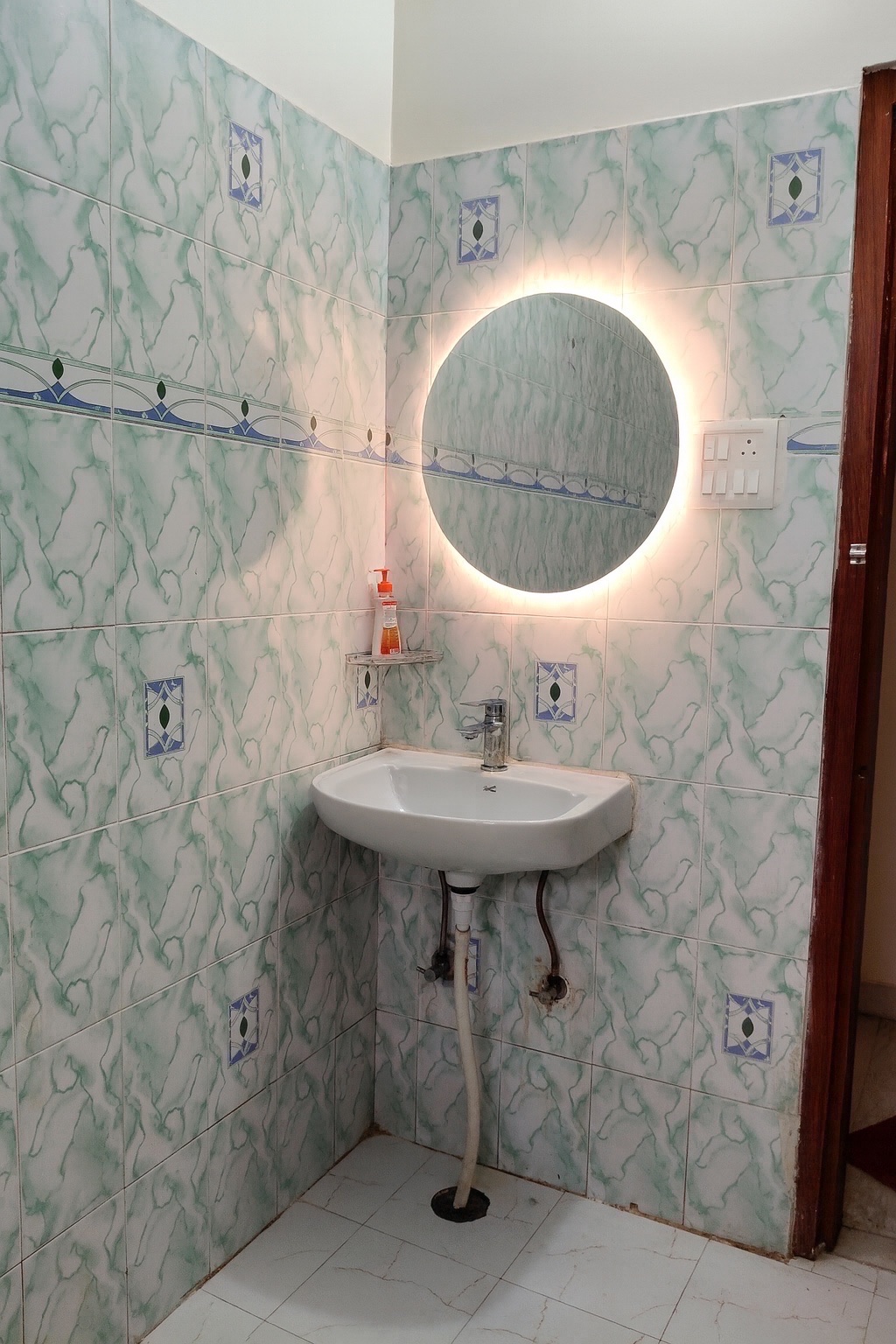 Pratham, Varanasi: washbasin under a backlit round mirror, green marbled tiling with a blue border and a handwash bottle on the corner shelf