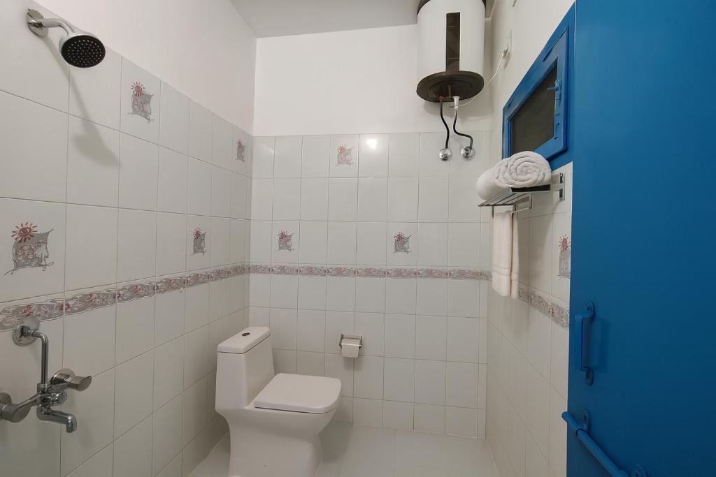 Santorini, Varanasi: white-tiled bathroom with a shower, western toilet, overhead water heater and rolled towels on the rail beside a blue door